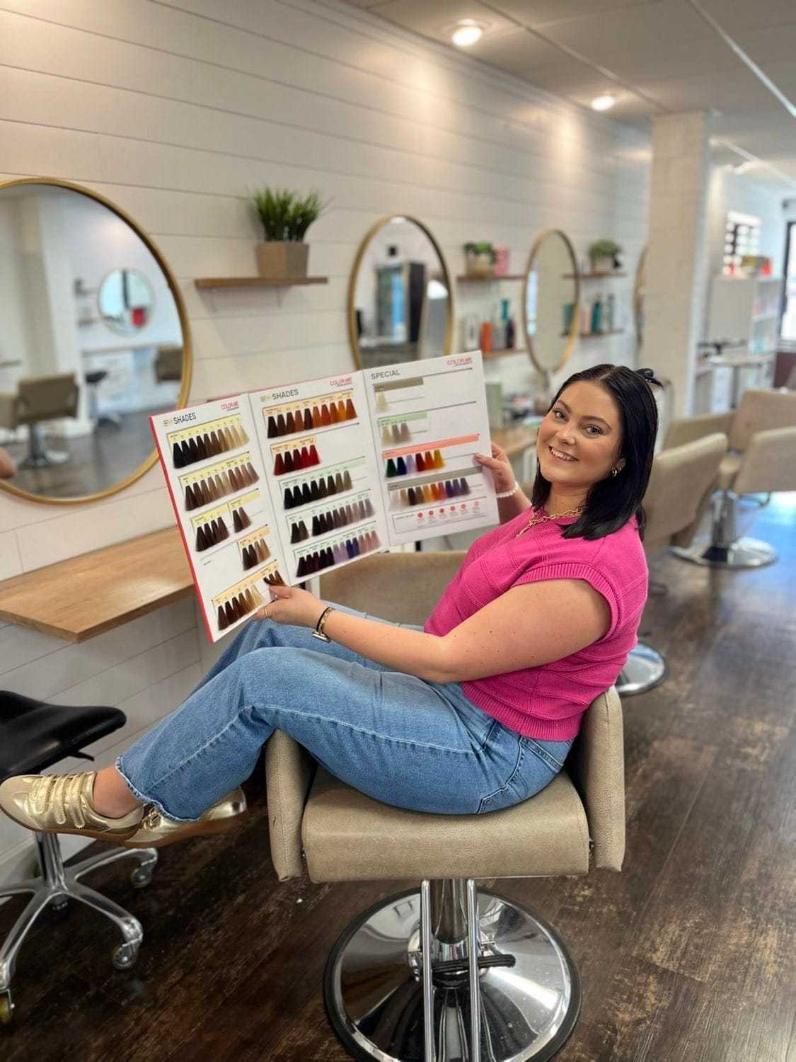 Woman in salon holding a hair color chart, seated on a styling chair.