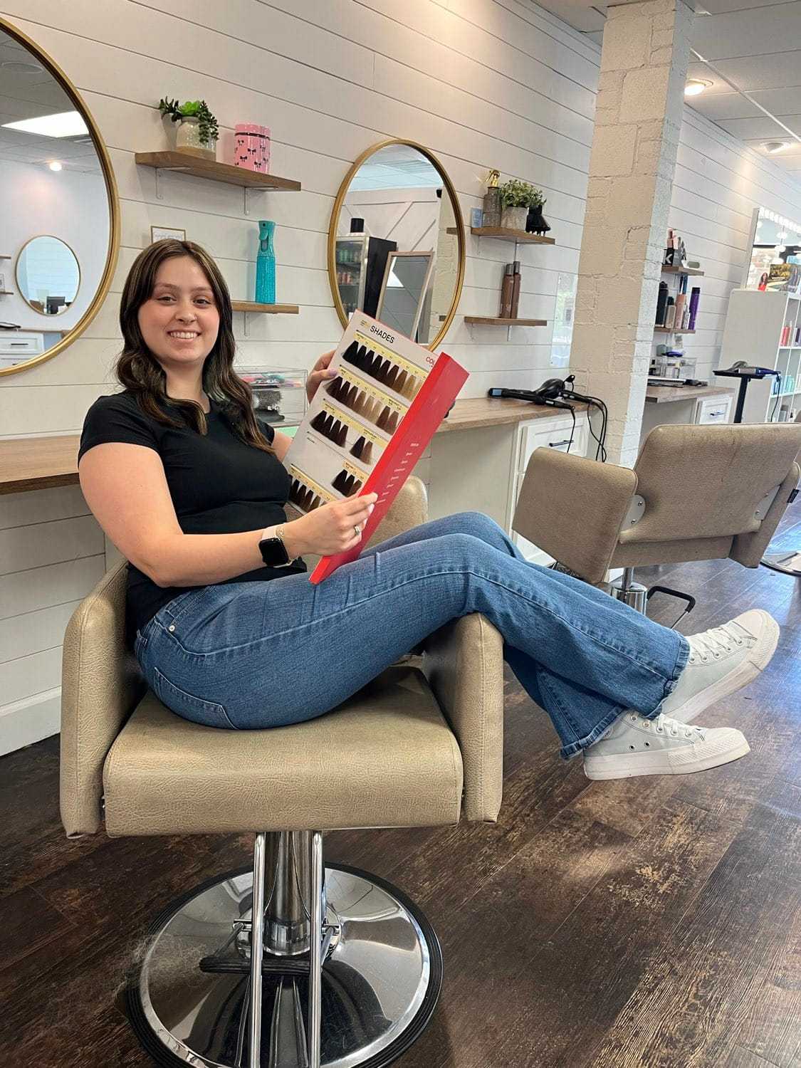 Woman sitting in salon chair holding a color chart, smiling, in a modern hair salon.